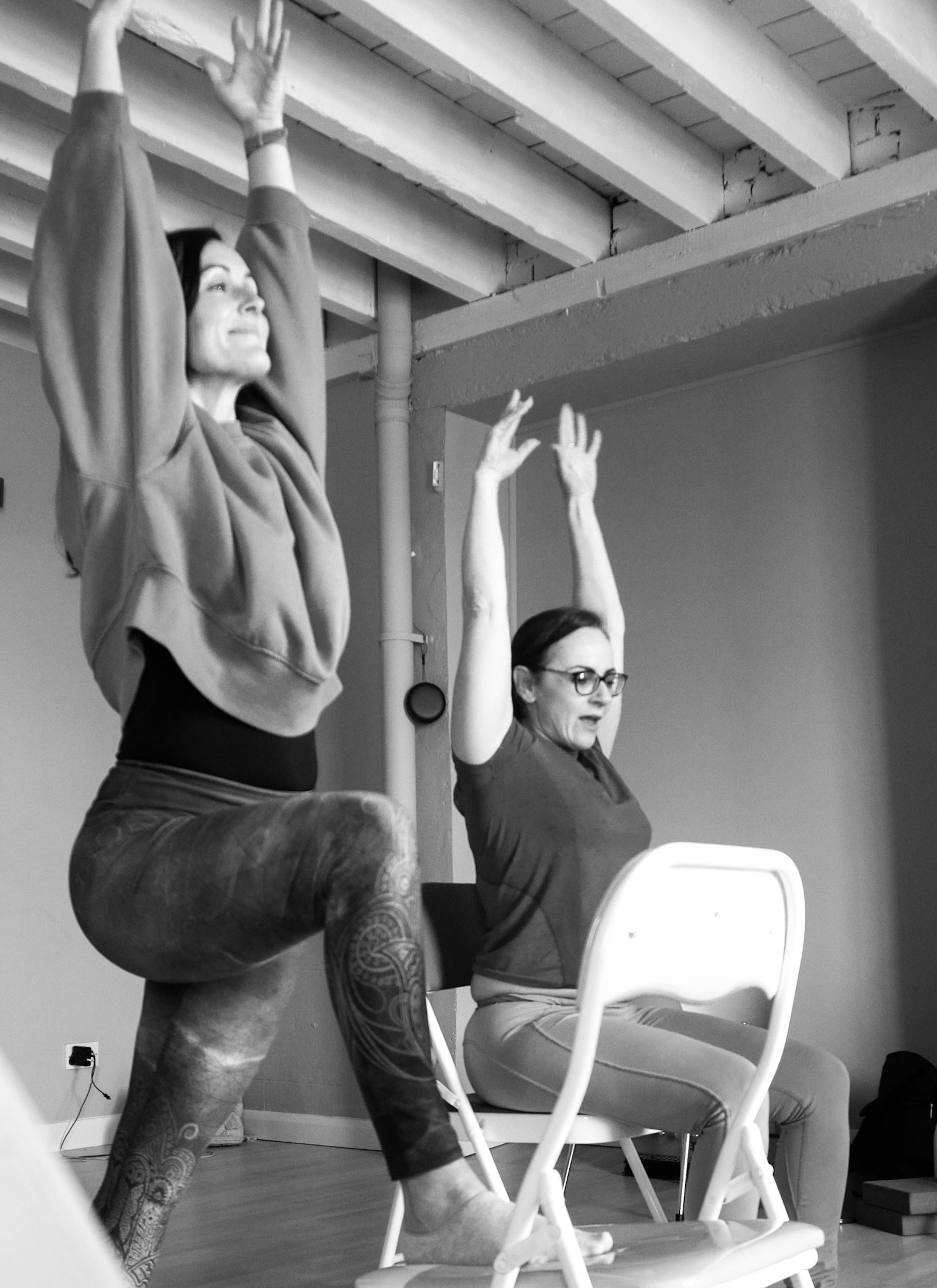 Two students using the chair as a prop for Warrior 1 posture on a yoga teacher training at the Shala