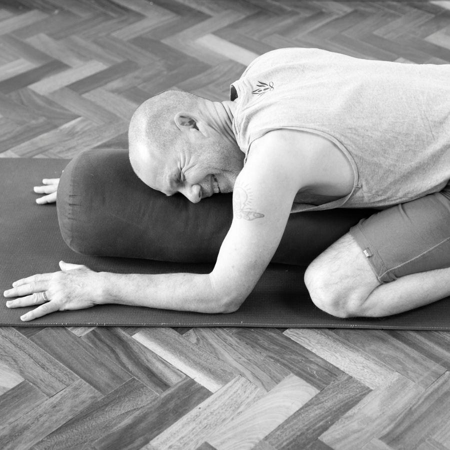Norman Blair practicing a restorative or Yin Yoga pose, commonly known as a supported Child's Pose, using a yoga bolster for support during his Yin Yoga Teacher Training at the shala